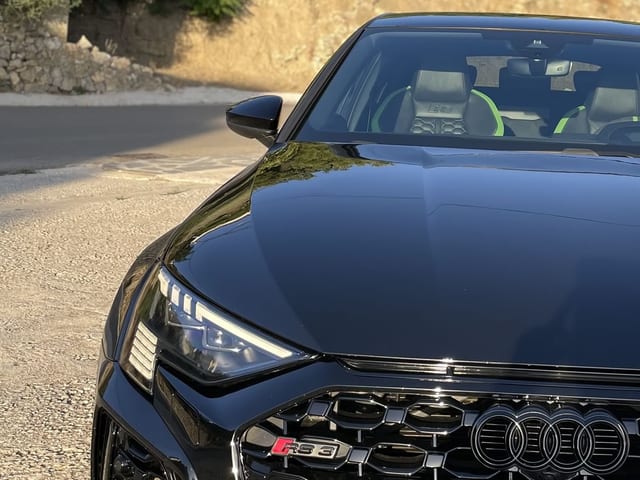 Vista frontal de un Audi RS 3 de color negro brillante en un entorno exterior, destacando la parrilla Singleframe, los aros de Audi y los faros LED detallados.